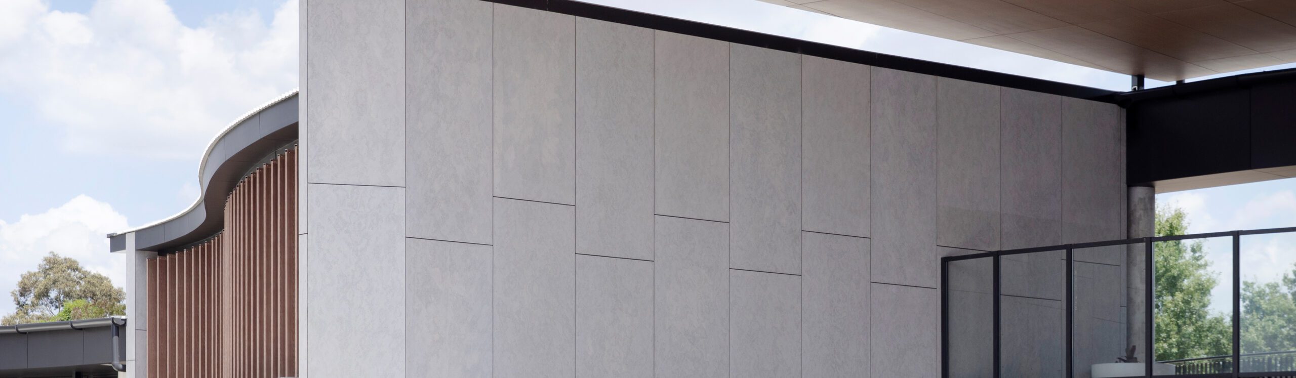 A modern building exterior with a large, minimalist white wall featuring subtle vertical and horizontal lines. To the right, there is a glass balcony and some greenery is visible in the distance. The edges of the roof and a partial view of the sky are also visible.