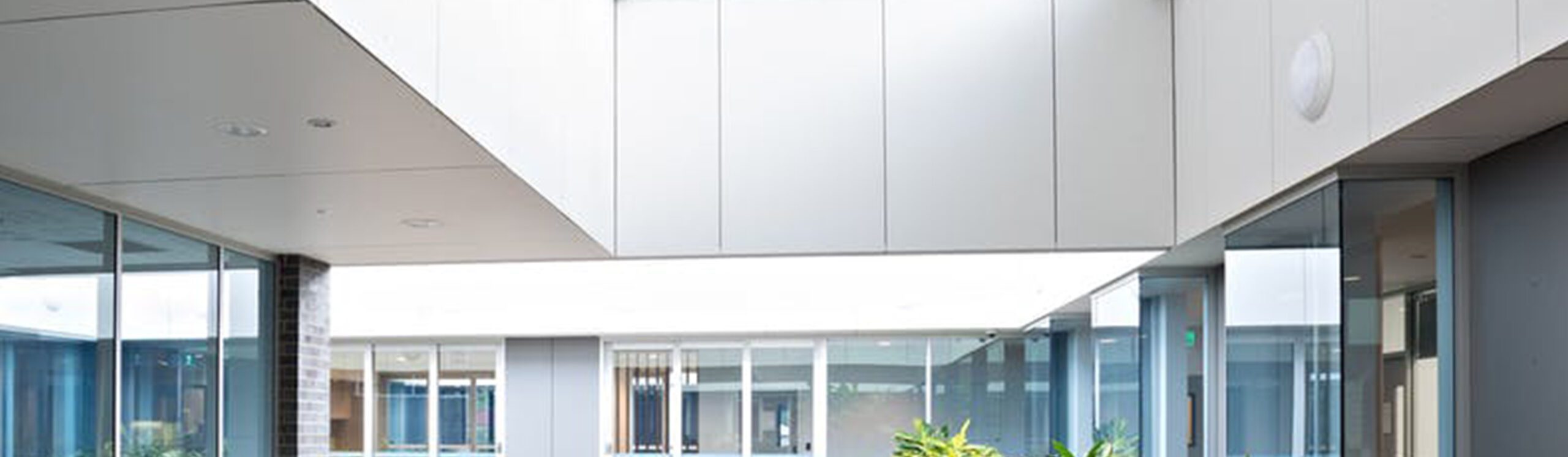 A modern indoor atrium with large windows allowing natural light. Clean lines, white ceilings, and sparse decoration create a minimalist aesthetic. A few green plants are placed throughout the space. Glass doors and windows line the perimeter, revealing adjacent rooms.