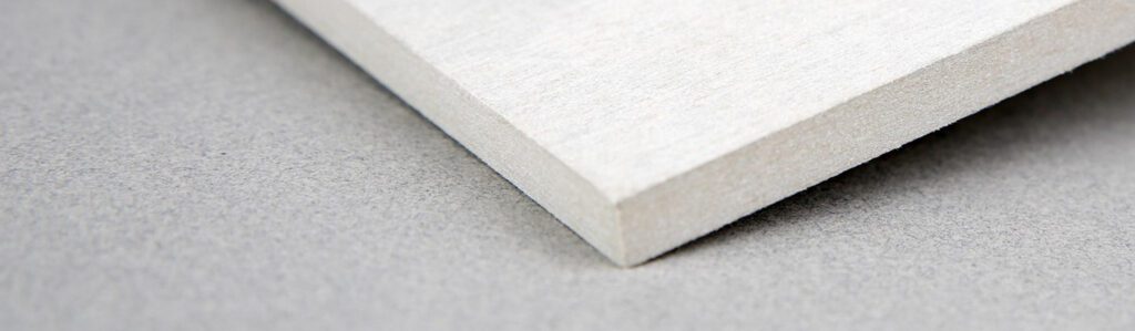 Close-up of the edge of a light-colored, rectangular piece of wood or fiberboard placed on a smooth, gray surface. The photo focuses on the corner of the board, showing its grain and texture.