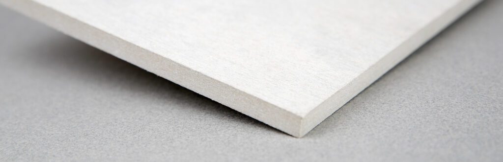 Close-up of the corner of a thin, white rectangular tile with a smooth surface. The tile lies on a slightly textured grey background. The focus is on the sharp edge and the thickness of the tile.