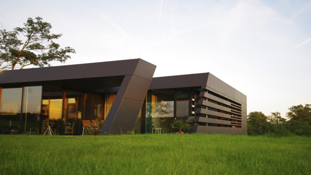 A modern house with sleek, angular architecture featuring large glass windows and dark exterior panels. The home is surrounded by a lush green lawn and trees, with a blue sky and light clouds in the background.