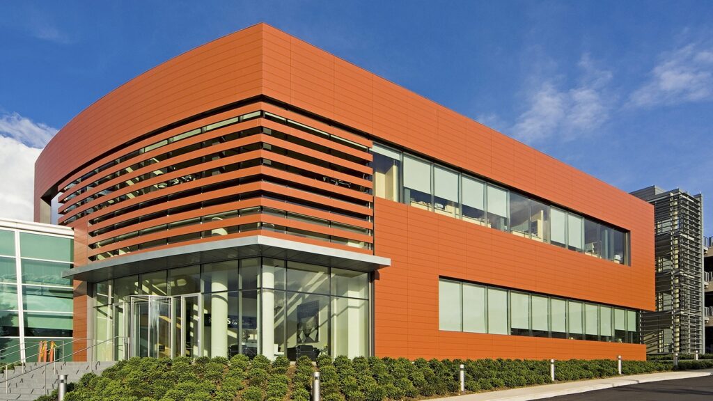 A modern, two-story building with red horizontal slats and large glass windows. The entrance features glass doors and stairs with metal railings. The landscaping includes small green shrubs around the building's perimeter. The sky is blue with scattered clouds.