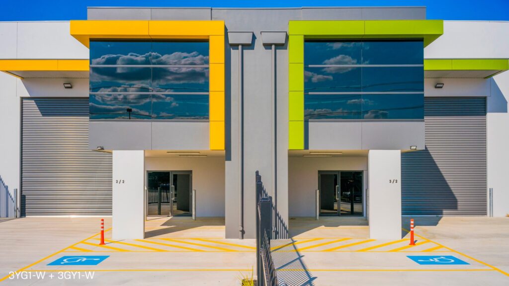 A modern commercial building with two mirrored units. The facade features large black-tinted windows, vertical yellow and green accents, and garage doors. There are designated parking spaces, including two handicapped spots, and a clear blue sky backdrop.